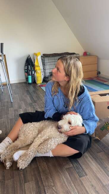 A young woman in a blue sweater sits on the floor, gently holding a fluffy white dog on her lap in a cozy, pet-friendly room.