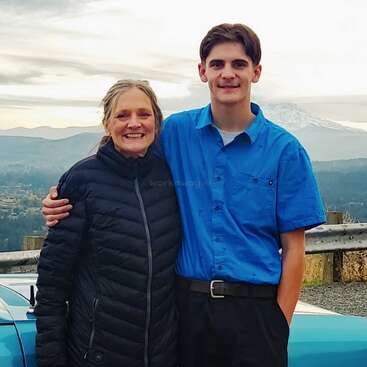 A smiling woman and young man stand arm-in-arm beside a blue car, with scenic mountains and cloudy sky visible in the background, enjoying a peaceful moment together.