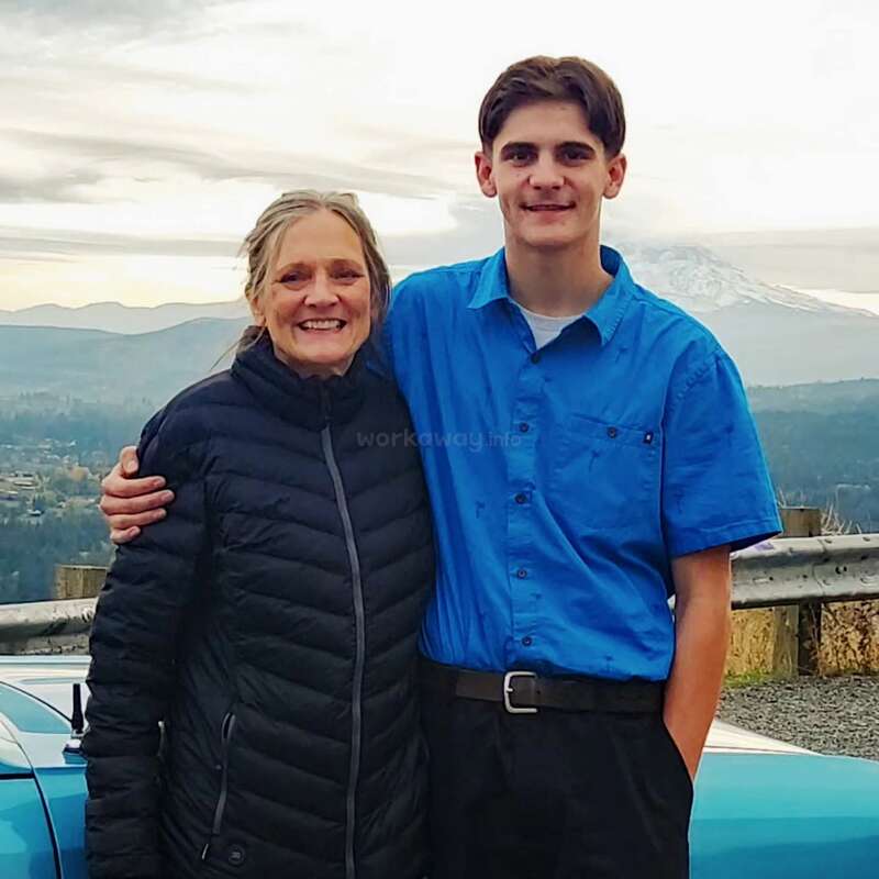 Une femme souriante et un jeune homme se tiennent bras dessus bras dessous à côté d'une voiture bleue, avec des montagnes pittoresques et un ciel nuageux visibles en arrière-plan, profitant d'un moment paisible ensemble.