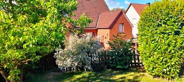 Un jardin ensoleillé avec des arbres et des buissons verdoyants se trouve devant une charmante maison en briques rouges avec un toit en tuiles rouges et une clôture en bois.
