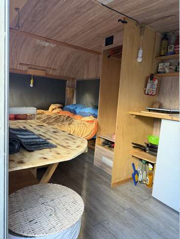This image shows the cozy interior of a camper with a wooden bed, small kitchen area, round wicker seat, shelves, and essential cooking supplies. Warm, rustic feel.