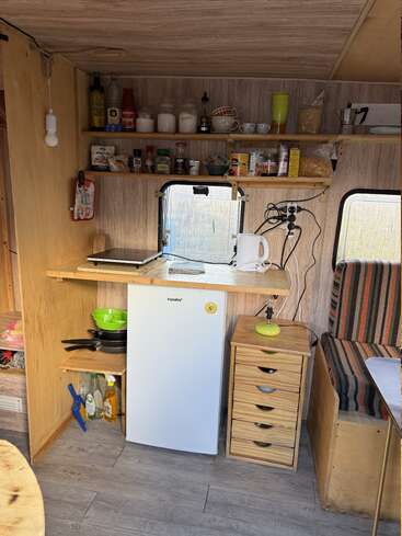 A compact camper kitchen with shelves holding various food items, a mini fridge, kettle, pans, cleaning supplies, and a small wooden drawer unit beside a bench.