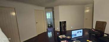 Ein geräumiger Raum mit dunklem Holzboden, drei geschlossenen Türen, einem sichtbaren Badezimmer, weißen Wänden, einem Fernseher, einem Tisch mit Papieren, einem Korbstuhl und einem kleinen Regal.