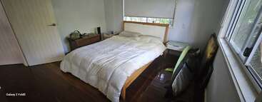 Ein gemütliches Schlafzimmer mit Holzboden, weißem Bett, Nachttisch und Jalousien. Kunstrahmen und ein grüner Stuhl stehen an der Wand. Minimalistische Einrichtung.