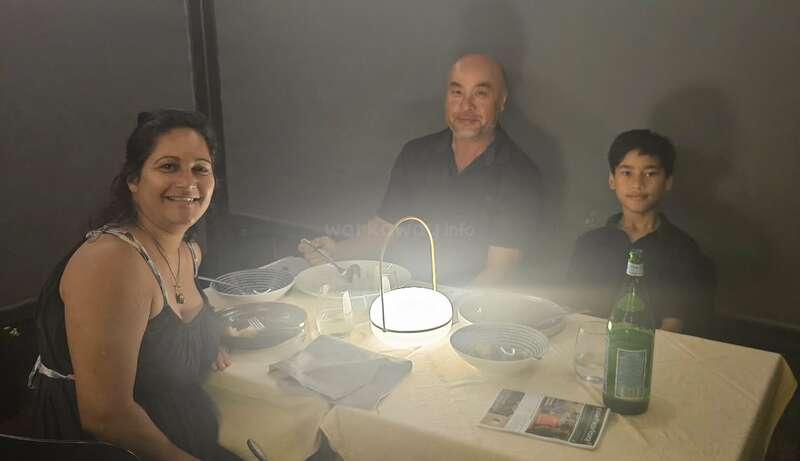 Eine dreiköpfige Familie sitzt lächelnd um einen kleinen Tisch mit leeren Schüsseln und Gläsern. In der Mitte steht eine helle Lampe, die eine warme Atmosphäre schafft.