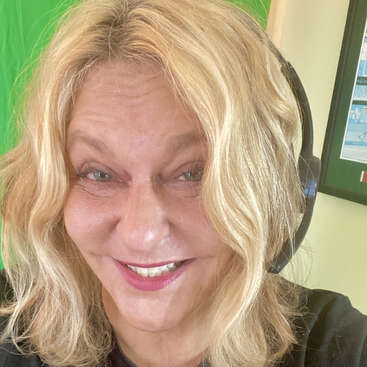 Une femme souriante aux cheveux blonds ondulés porte un casque audio. Elle se trouve à l'intérieur, devant un fond vert, et semble s'amuser.