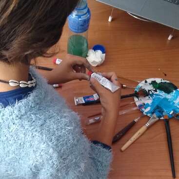 Das Bild zeigt eine Frau, die mit einem blauen Pullover und einer Muschelkette bekleidet ist und mit dem Rücken zur Kamera auf einem Holztisch steht, umgeben von Malutensilien.