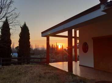 A beautiful sunset casts warm light on a modern house with a tiled porch, surrounded by tall cypress trees, rustic fencing, and a peaceful grassy yard.