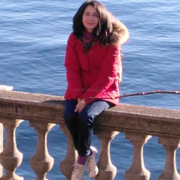 Eine Frau in einem roten Mantel sitzt auf einer Steinbrüstung am Wasser. Sie lächelt, hat dunkles Haar und trägt Jeans und Turnschuhe.
