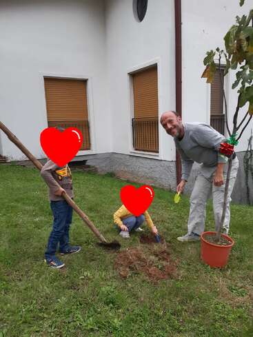 A man and two children, faces covered with red hearts, are digging in a garden near a house, preparing to plant a small tree in a pot.