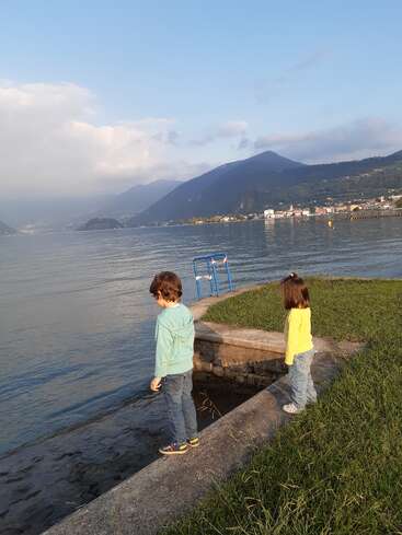 Two children stand by a calm lakeshore, gazing at the water. Mountains and a town are visible across the lake under a partly cloudy sky. Peaceful scene.