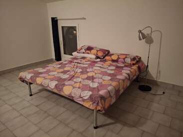 A minimalist bedroom features a double bed with colorful polka-dot bedding, two pillows, a small window with dark curtains, tiled floor, and a metal floor lamp.