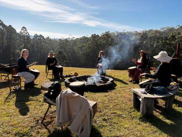 Cinco pessoas sentadas em círculo ao redor de uma fogueira enfumaçada, tocando tambores, ao ar livre em um dia ensolarado, cercadas por grama verde e árvores altas. Atmosfera tranquila.