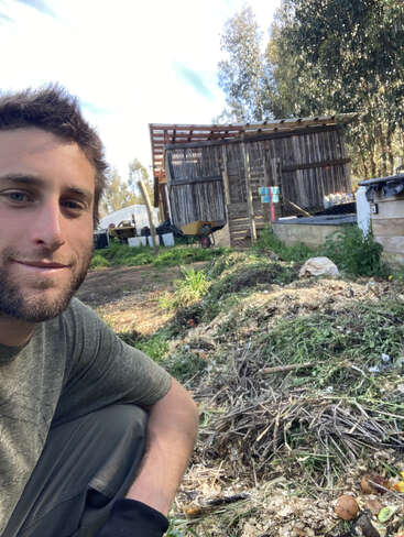 A man smiles at the camera while crouching beside a compost pile. Behind him is a rustic wooden shed, wheelbarrow, greenery, and trees under a bright sky.