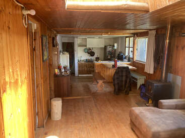 This cozy wooden cabin interior features a kitchen with hanging pots, a dining bench, wood stove, large windows, a sofa, rustic decor, and natural sunlight.