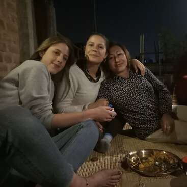 Three women sit closely together on a mat at night, smiling, enjoying each other's company. There is a metal plate of food and a glass nearby.