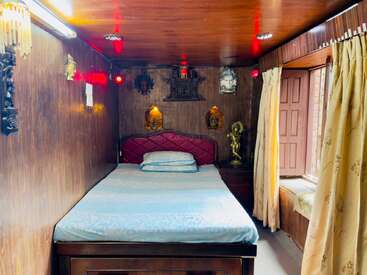 A cozy wooden bedroom with a double bed, red headboard, decorative statues, wall art, warm lighting, yellow curtains, and a large window seat area.