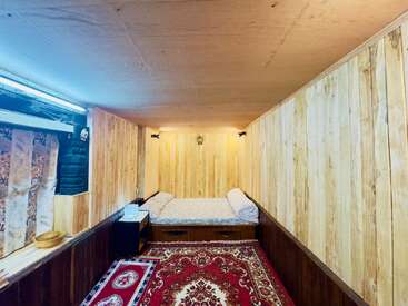This cozy wooden room features a single bed, ornate red carpet, bedside table, towels, soft lighting, window with decorative curtains, and a warm, rustic ambiance.