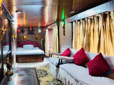 This cozy room features a wooden ceiling, golden curtains, a bed, a sofa with red cushions, ornate statues, soft lighting, and decorative rugs and art.