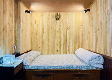 This image shows a cozy wooden room with a neatly made bed, soft pillows, rolled blanket, side table, wall lights, and a decorative wall piece above.
