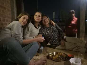 Drei Frauen sitzen nachts eng beieinander auf einer Matte, lächeln und genießen die Gesellschaft der anderen. Daneben stehen ein Tablett mit Essen und ein Glas.