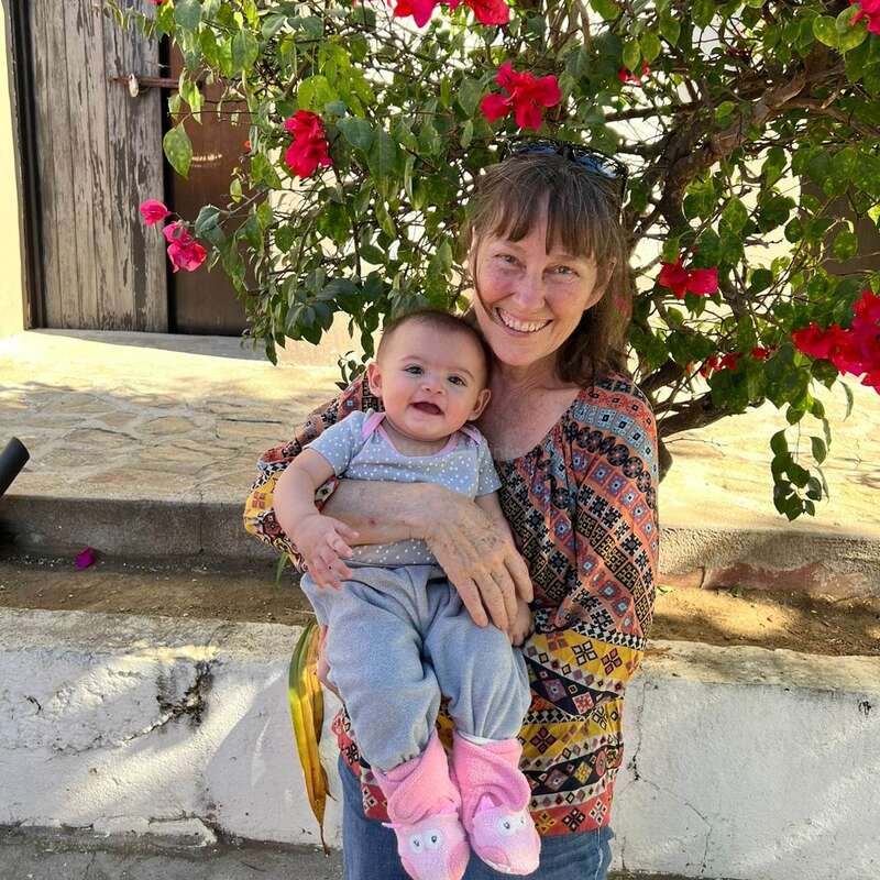 La imagen muestra a una mujer con un bebé en brazos delante de un arbusto en flor. La mujer lleva una blusa estampada y el bebé viste un atuendo gris y rosa.