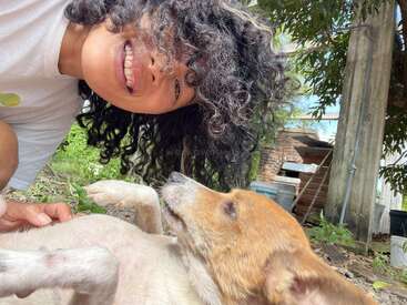 A smiling person with curly hair bends down to pet a brown and white dog lying playfully on the ground outdoors, surrounded by greenery and rustic structures.