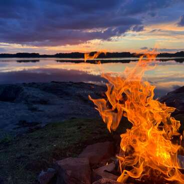 Das Bild zeigt eine ruhige Seeszene bei Sonnenuntergang, mit einem brennenden Lagerfeuer im Vordergrund und einem bewölkten Himmel, der sich auf der Wasseroberfläche spiegelt.