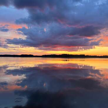 La imagen representa una serena escena lacustre al atardecer, con un cielo vibrante con nubes, una tranquila masa de agua y una lejana línea de árboles, todo ello reflejado en las tranquilas aguas.