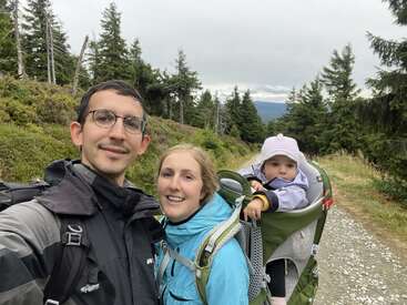 A family of three, dressed in outdoor gear, enjoys hiking on a forest trail. The baby is carried in a backpack, surrounded by tall trees.