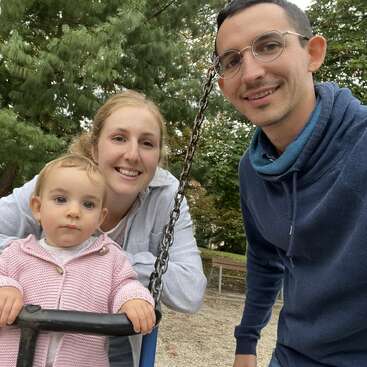 A family photo outdoors shows a smiling man and woman beside a young child on a swing. Trees and benches create a peaceful, natural park background.