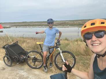 A man and woman in helmets are cycling outdoors. The man is with a bike and child trailer, beside a marsh, while the woman takes a selfie.