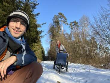 Two people enjoy a snowy walk in a forest. One person smiles at the camera, while the other pushes a stroller along the snow-covered path.