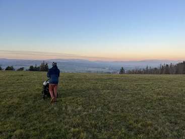 A person pushes a stroller across a vast grassy field at sunset. Rolling hills, distant trees, and a soft pastel sky create a peaceful scene.