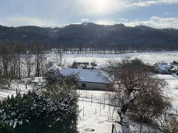 La imagen muestra un pintoresco paisaje rural nevado con colinas al fondo, árboles, casas y campos cubiertos de nieve bajo un cielo luminoso y soleado.