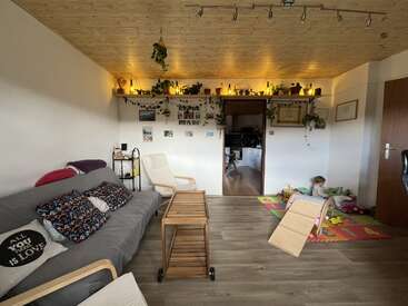 Acogedora sala de estar con techo de madera, sofá, sillón, niño juguetón sobre alfombra de colores, carrito de madera, plantas en macetas sobre estantes y cálida iluminación ambiental en toda la estancia.