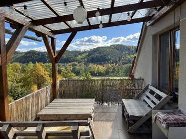 Una acogedora terraza de madera con una mesa y un banco de palets con vistas a un pintoresco paisaje de verdes colinas y árboles bajo un cielo azul con nubes. Ambiente relajante.