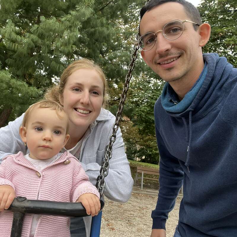 A family photo outdoors shows a smiling man and woman beside a young child on a swing. Trees and benches create a peaceful, natural park background.