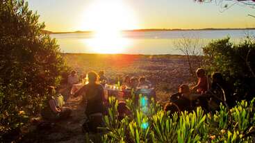 Eine Gruppe von Menschen genießt bei Sonnenuntergang ein entspanntes Picknick im Freien am Wasser, umgeben von Grün, mit gemeinsamen Essen, Gesprächen und der friedlichen Naturkulisse.