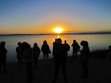 Eine Gruppe von Menschen steht am Ufer und hebt sich von einem farbenfrohen Sonnenuntergang über dem ruhigen Wasser ab, was eine ruhige und friedliche Atmosphäre in der Abenddämmerung schafft.