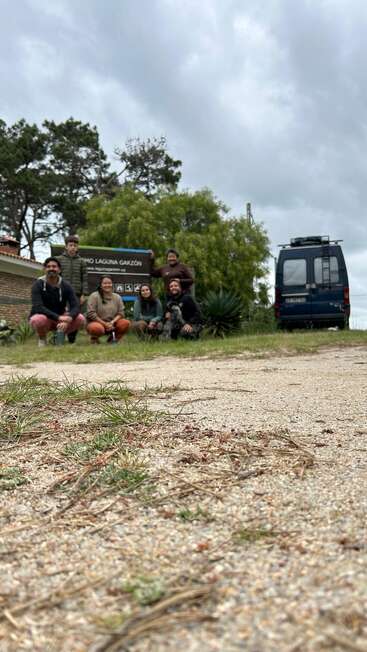Eine Gruppe von sechs Personen posiert vor einem Schild mit der Aufschrift "Laguna Garzón". Es gibt einen blauen Lieferwagen und grüne Bäume unter einem bewölkten Himmel.