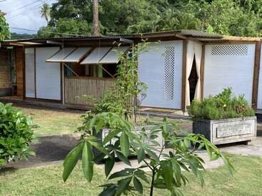 The image depicts a small, white building with a wooden frame, featuring a covered porch and a garden area with plants and trees in the foreground.