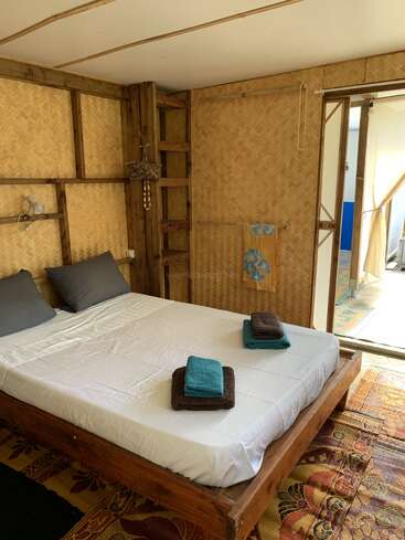 The image depicts a serene bedroom with a wooden bed frame, white sheets, and a woven bamboo wall, featuring a doorway to an outdoor area with a rug and curtains.