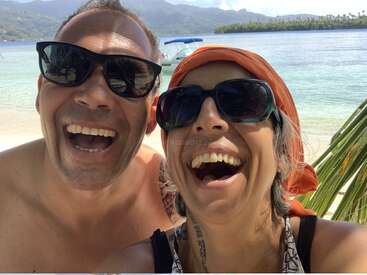 The image shows a man and woman taking a selfie on a beach, with a boat and palm trees in the background, wearing sunglasses and smiling.