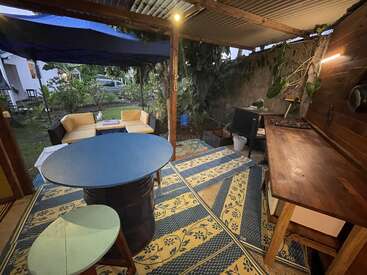 This cozy outdoor kitchen features a wooden countertop, small stove, round blue table, patterned rug, yellow sofas, and plants, creating a relaxed and inviting atmosphere.