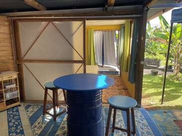 A cozy, rustic room with wooden walls and sliding doors opens to a garden. A blue barrel table and two stools sit on patterned rugs.