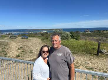 A couple stands by a railing on a sunny day. Behind them are grassy fields, picnic tables, and a scenic ocean view with boats and a house.