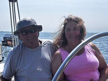 A man and woman are smiling, enjoying a sunny day on a boat at sea. The woman’s hair blows in the wind, evoking joy and relaxation.