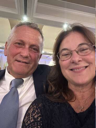 Un homme souriant en costume-cravate est assis à côté d'une femme portant des lunettes et une robe en dentelle. Ils ont l'air heureux, probablement en train d'assister ensemble à un événement officiel.