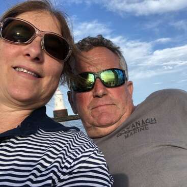 The image shows a selfie of a man and woman posing together outdoors, with a lighthouse visible in the background on a sunny day.
